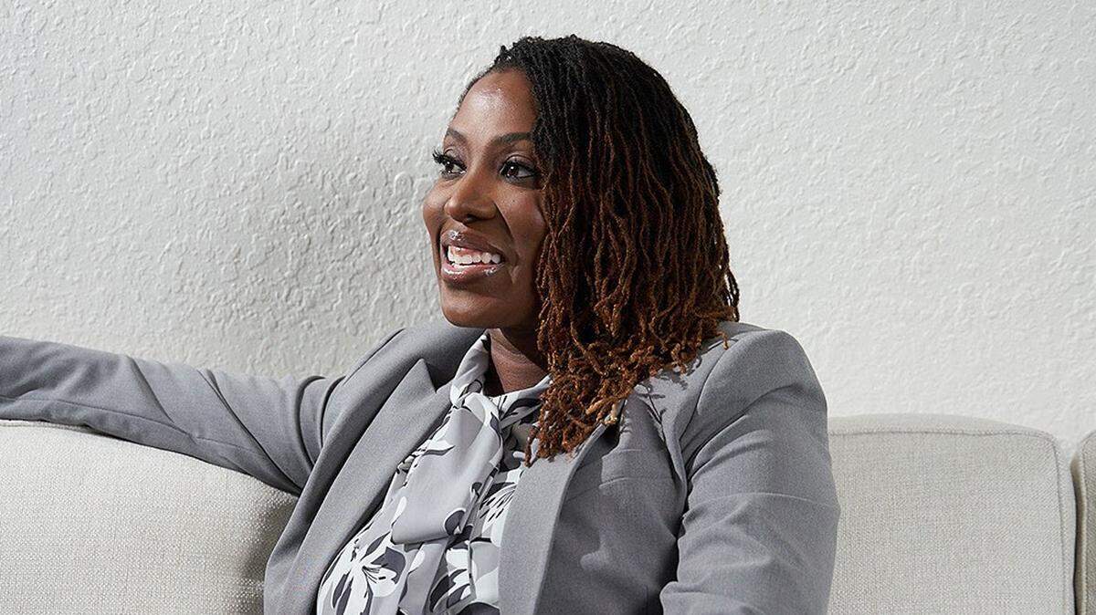 A Black woman in a grey suit on a grey couch smiles, looking off-camera.