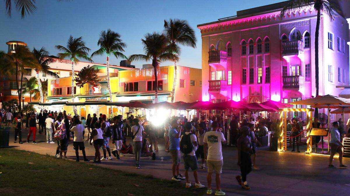 Large groups of people gathering outside bars and clubs in Miami Beach with palm trees in the background.