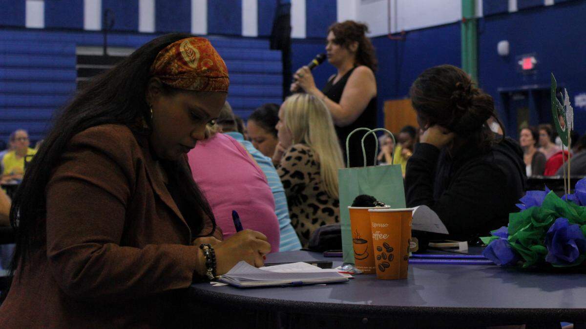 A young Black woman with an orange headband and brown suit takes notes with a pen and a pad of paper.