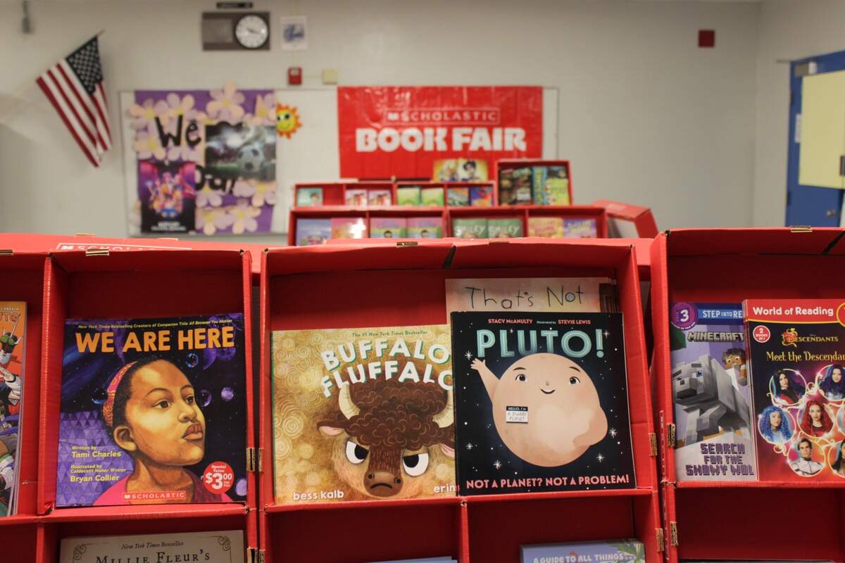 An image of a bright red book display with children’s books “We Are Here,” “Buffalo Fluffalo,” and “Pluto: Not a Planet, Not a Problem,” with the Scholastic book fair baner in the background.