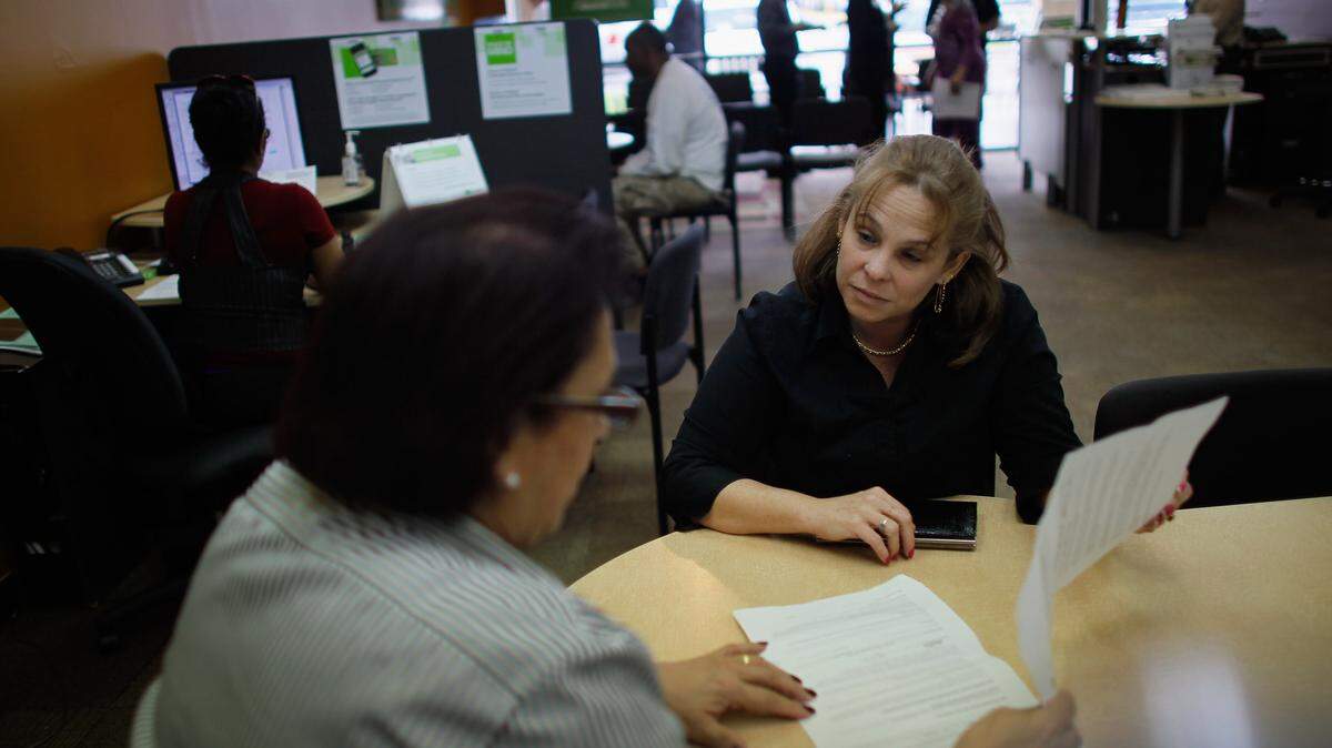 Two women sit across from each other at a table looking at paperwork together.
