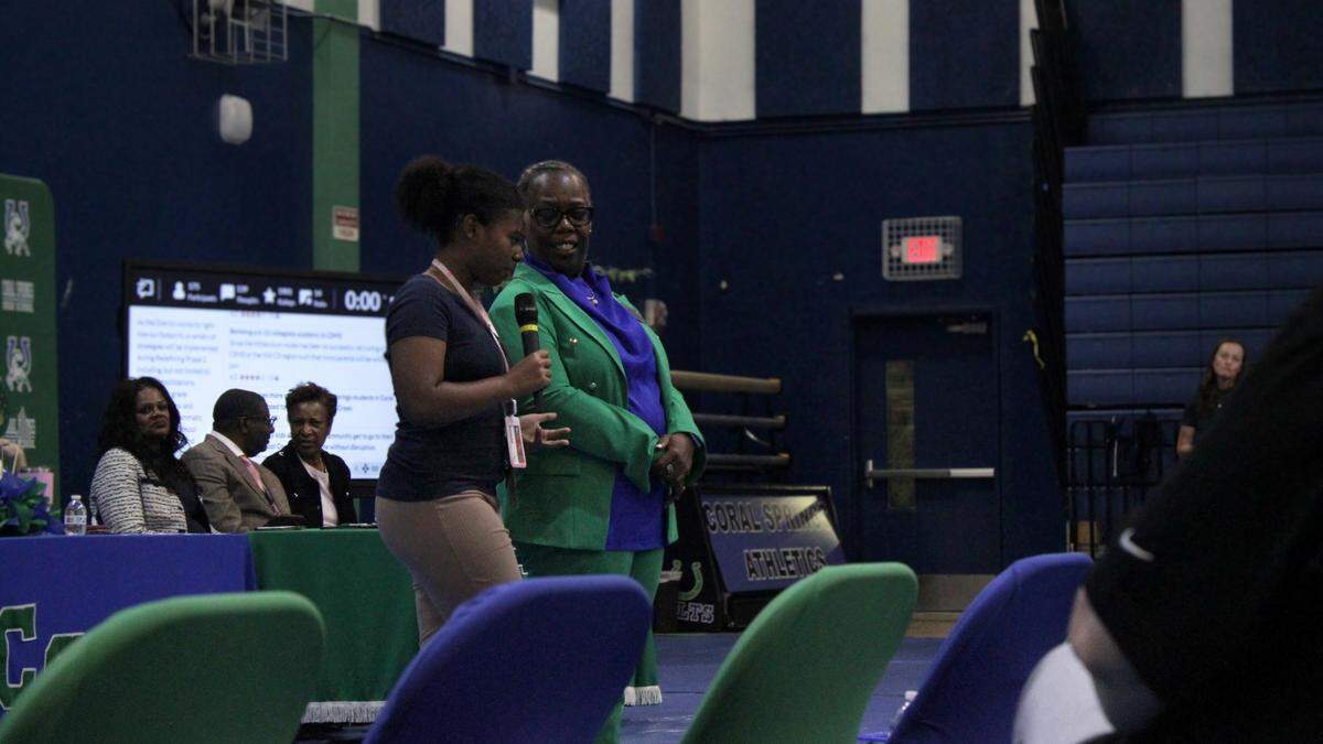 A young Black girl speaks into a microphone, standing next to a smiling older Black woman in a bright green pantsuit.