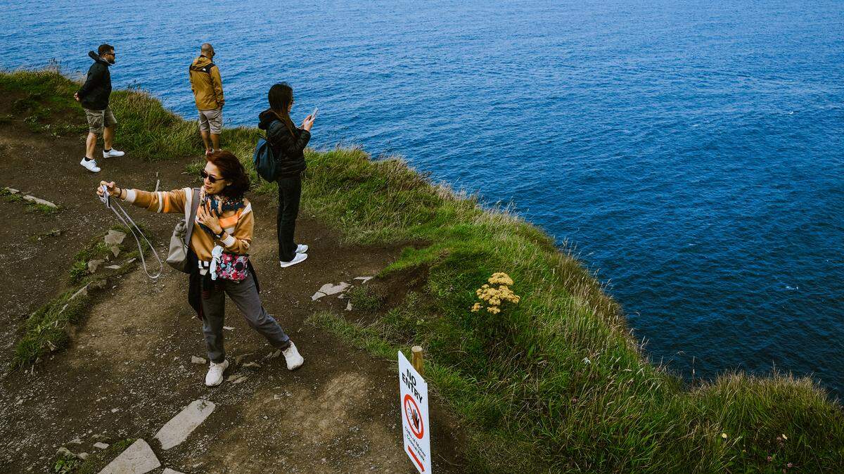 A group of four tourists lean over and take photos and selfies at the edge of the Cliffs of Moher.
