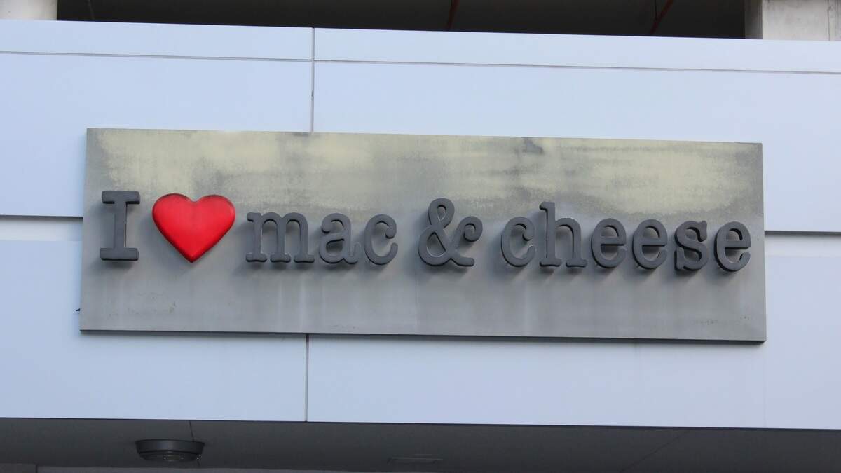 Photo with business sign for I Heart Mac and Cheese. Black typewriter font, red heart, on industrial-style grey backing.
