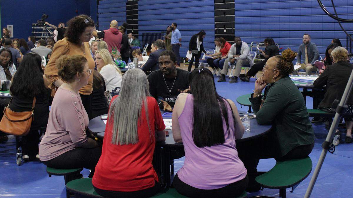 Five adults sit around a circular table in a blue high school gymnasium.