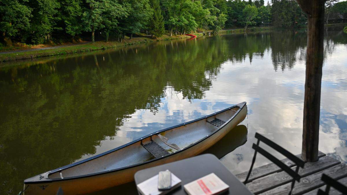 View of the terrace of a cabin on the water, with a book and a laptop on a table, a canoe nearby, and the lake in the background.