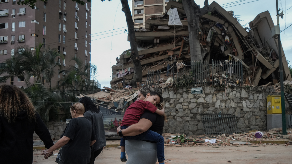 Women and a young child walk out of the frame, set against the backdrop of a partially-collapsed apartment building.