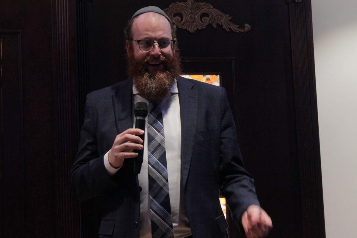 Rabbi Yankie Denburg smiles while speaking onstage. He is holiding a microphone and wearing a blue suit with a grey yarmulke.