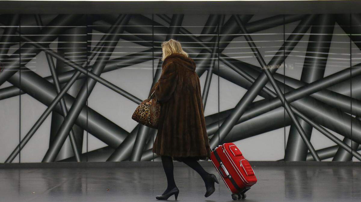 A woman wearing a fur coat carries a handbag and drags a suitcase behind her