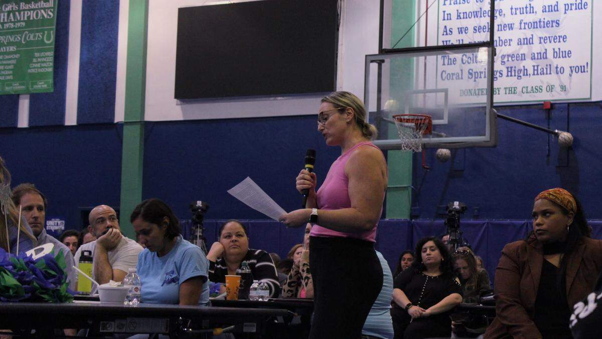 A blonde woman in a pink top speaks into a wireless microphone while holding a sheet of paper.