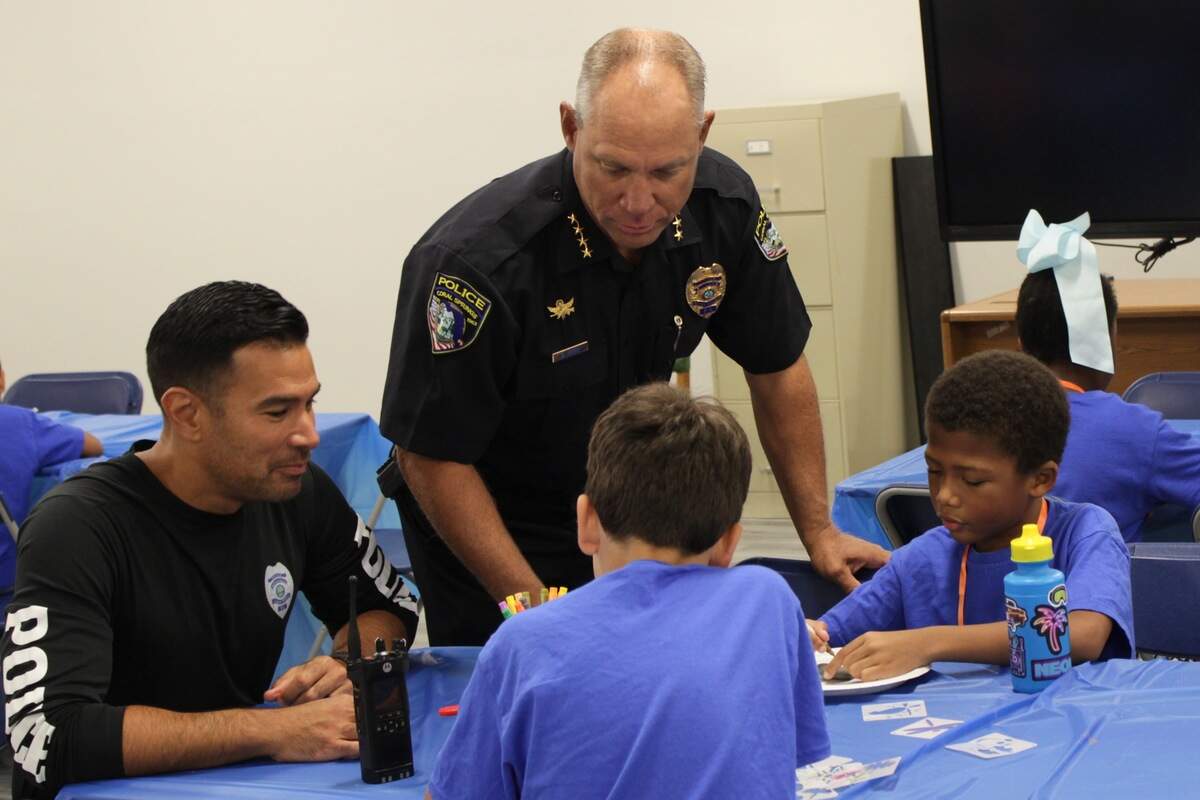 Two police officers and two children sit at a table with a bright blue tablecloth. Chief Brad Mock, center, talks to the children, who are focused on painting palm-sized stones.