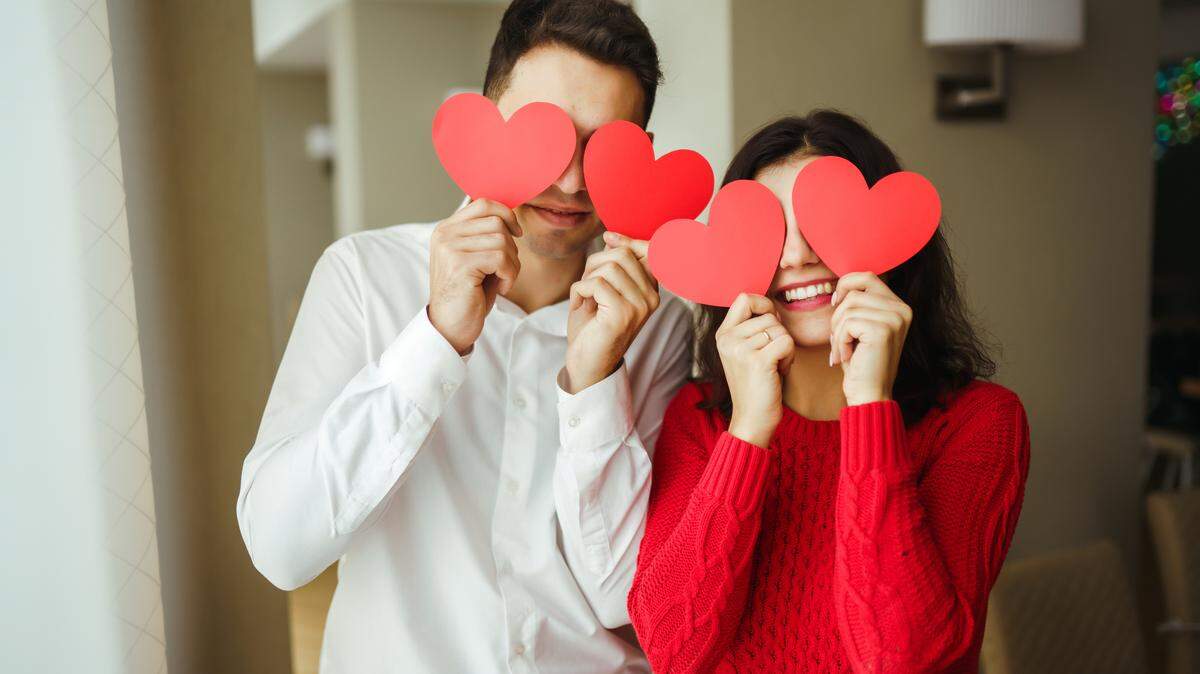 Young cheerful couple in love holding red paper hearts over eyes and smiling. Man wearing white button down shirt, woman wearing red sweater.